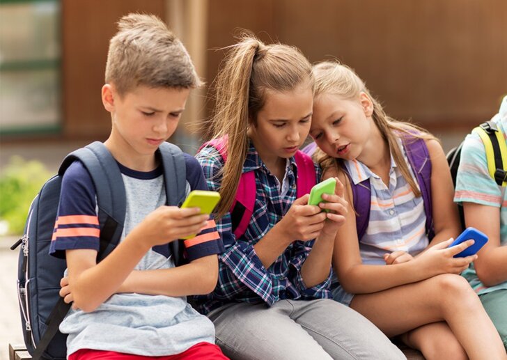 Schulkinder sitzen nebeneinander und schauen ins Handy | © iStock/dolgachov